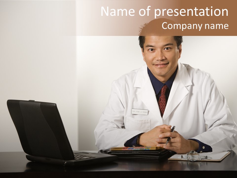 Asian American Male Doctor Sitting At Desk With Charts And Laptop Computer Looking At Viewer. PowerPoint Template