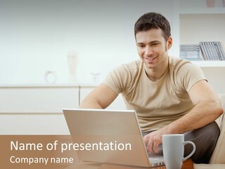 Happy Young Man In T-Shirt Sitting On Sofa At Home, Working On Laptop Computer, Smiling. PowerPoint Template