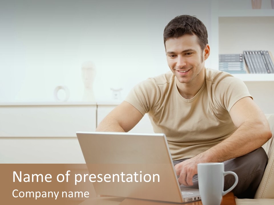 Happy Young Man In T-Shirt Sitting On Sofa At Home, Working On Laptop Computer, Smiling. PowerPoint Template