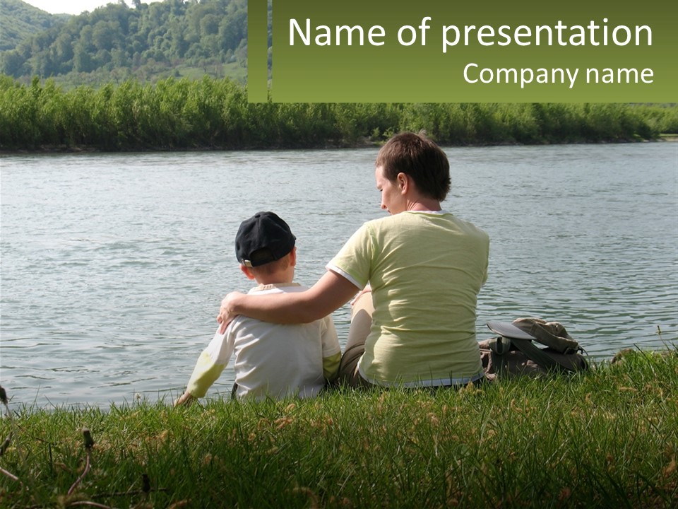 A Man And A Child Sitting On The Grass Near A Body Of Water PowerPoint Template